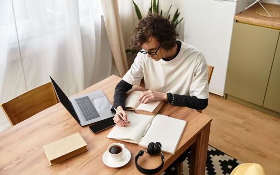 A person with curly hair sits at a wooden table, writing in a notebook. A laptop, a closed book, a cup of coffee, and headphones are also on the table. The room has wooden flooring and a patterned rug.