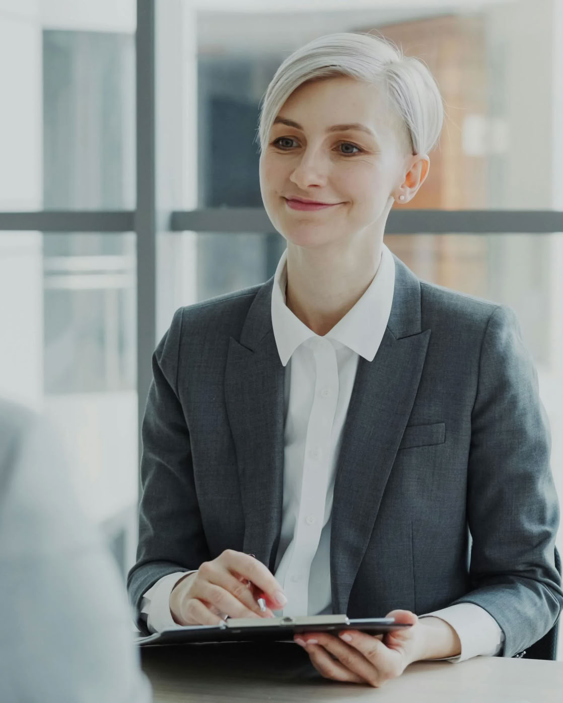 A person with short hair in a suit holding a clipboard, sitting across from another person in an office setting.