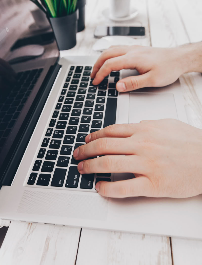 Hands typing on a laptop keyboard on a white desk with a plant, smartphone, and cup in the background.