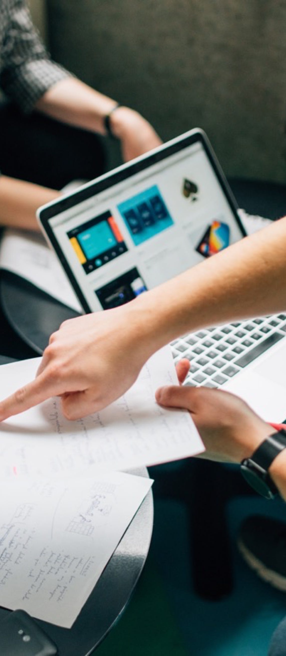 two people sat at a desk with a laptop and one person is pointing at paperwork