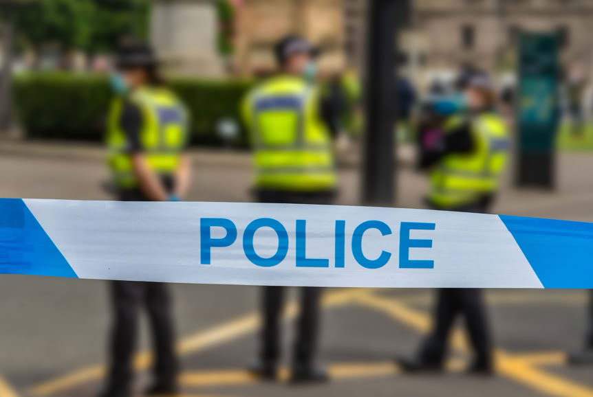 A police tape with the word "POLICE" in blue letters is in the foreground, with three uniformed officers standing in the blurred background.