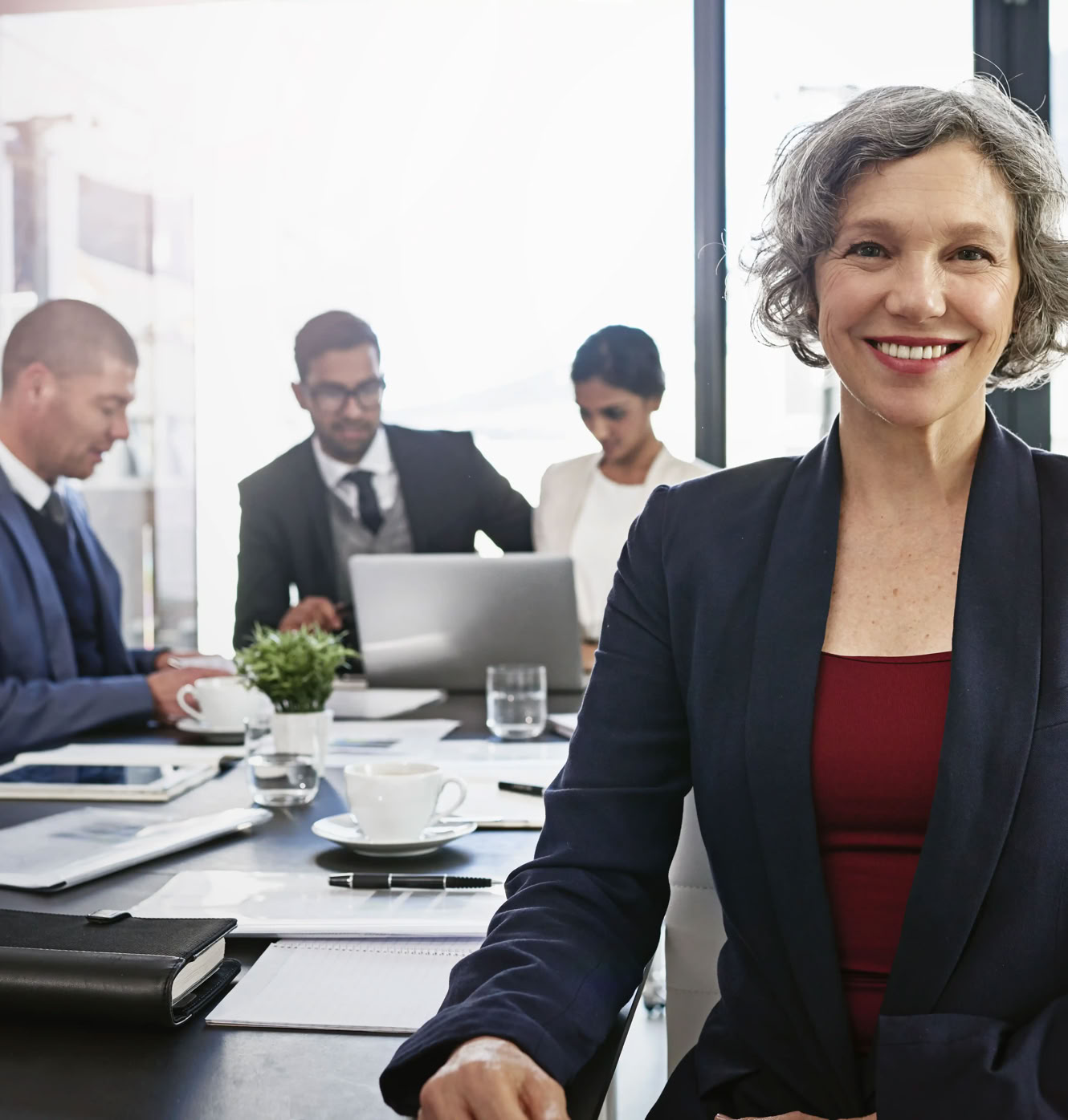 A woman in a suit smiling at the camera, with three people in business attire working at a table in the background.