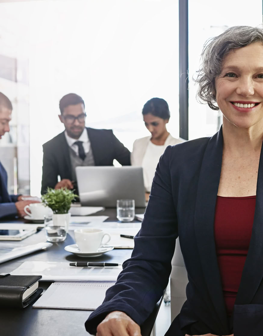 A woman in a suit smiling at the camera, with three people in business attire working at a table in the background.