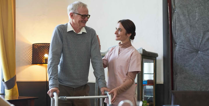 An elderly person using a walker is assisted by a caregiver in a room with a table, lamp, and curtains.