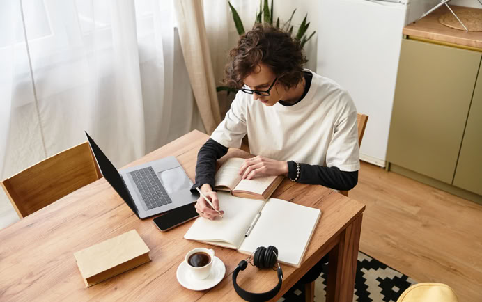 A person with curly hair sits at a wooden table, writing in a notebook. A laptop, a closed book, a cup of coffee, and headphones are also on the table. The room has wooden flooring and a patterned rug.